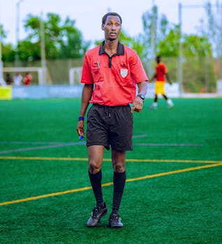 A soccer referee in red uniform on a green field during a match. Outdoor setting.