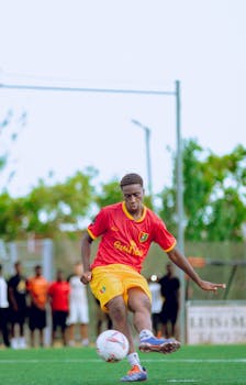 A young soccer player in a red and yellow uniform kicking a ball outdoors.