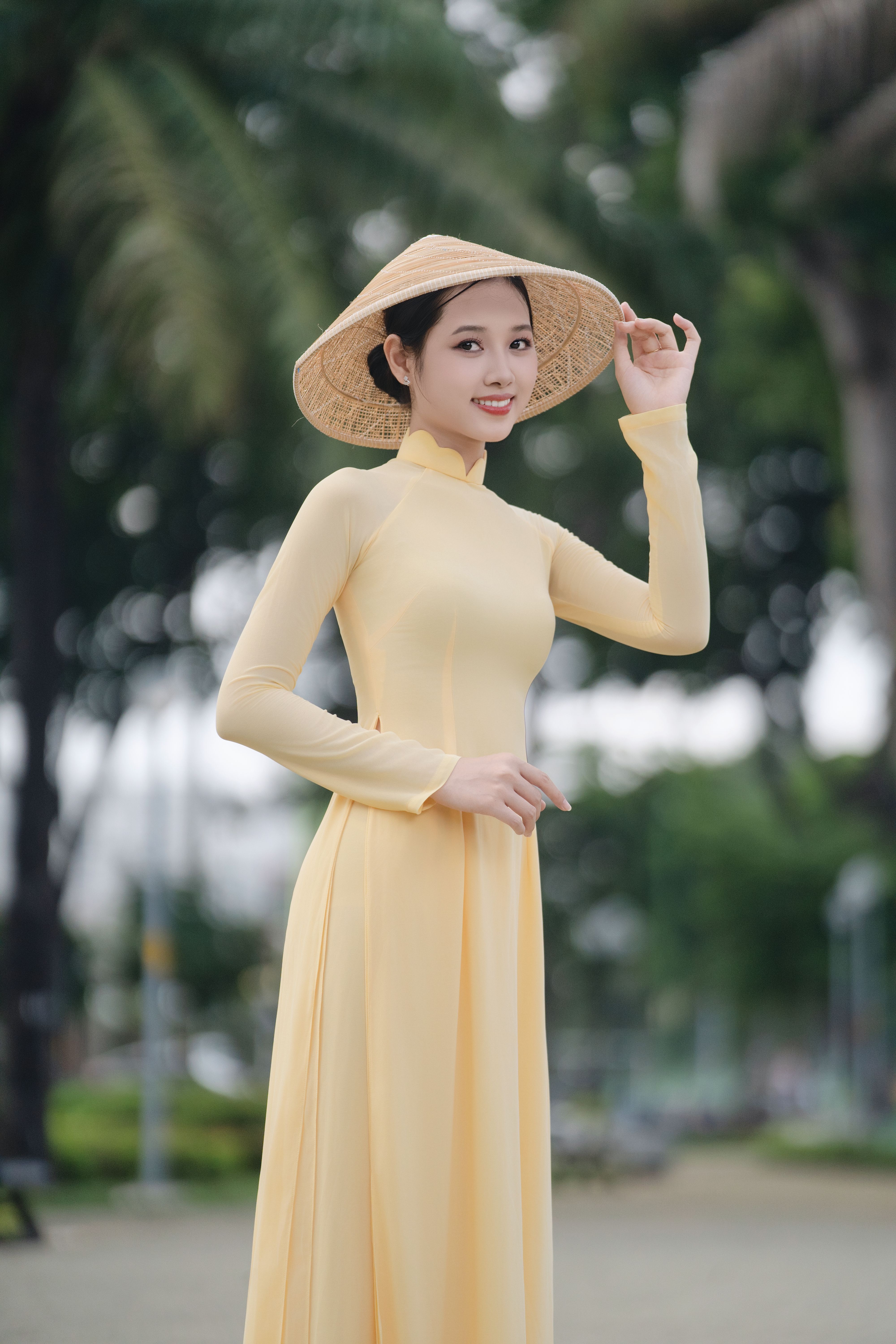 Young woman in yellow Ao Dai with conical hat, posing outdoors.