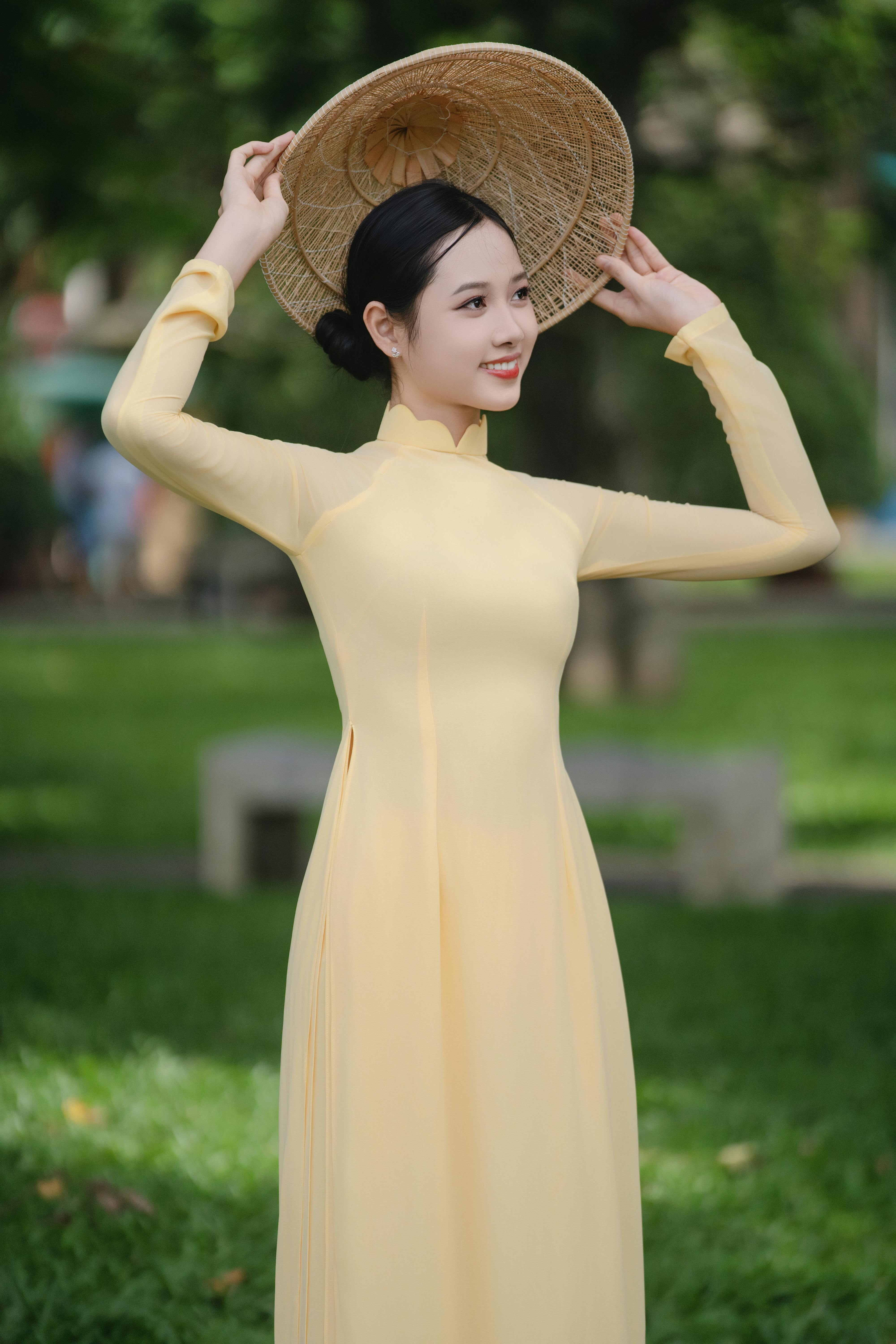 Young woman in a yellow ao dai holding a conical hat in a sunny park setting.