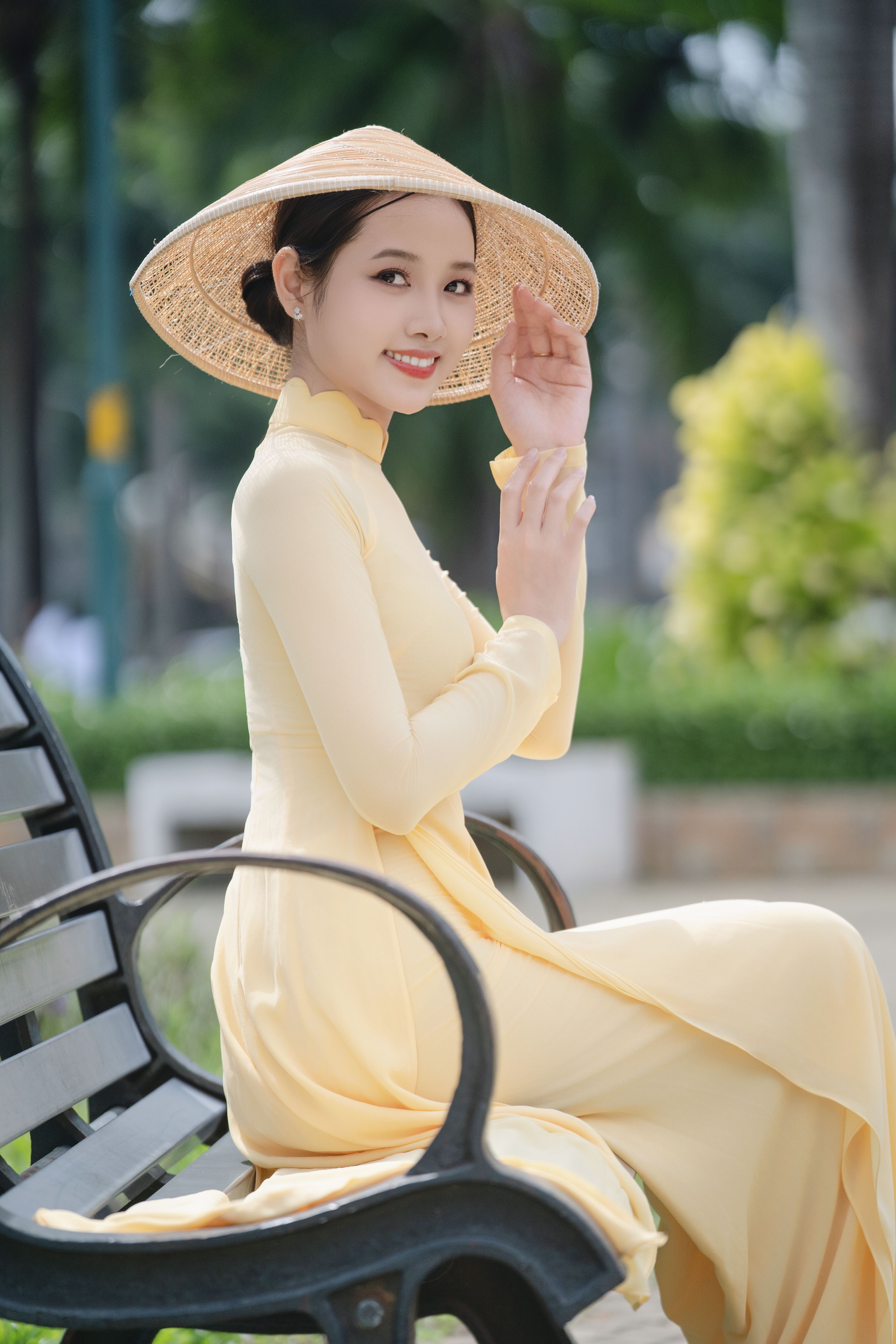 A graceful woman wearing a yellow Ao Dai and conical hat sitting on a park bench, symbolizing traditional Vietnamese culture.