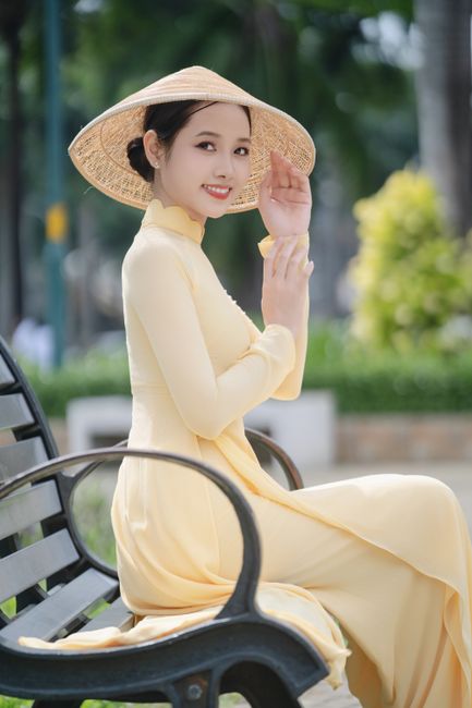 A graceful woman wearing a yellow Ao Dai and conical hat sitting on a park bench, symbolizing traditional Vietnamese culture.
