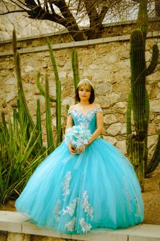 A young girl in a vibrant blue quinceañera dress posing by cacti and stone walls outdoors.