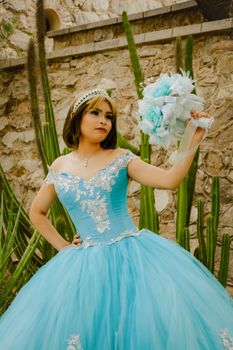 A young woman in a blue quinceañera dress poses elegantly outdoors with a bouquet.