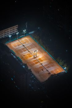 Illuminated tennis court viewed at night from above, located in a city setting.