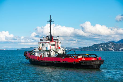 A vibrant red tugboat navigates the calm blue sea under a clear sky with clouds.