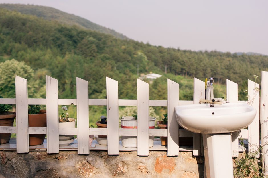 A tranquil outdoor scene featuring a rustic sink against a white fence with potted plants, set in a lush green landscape.