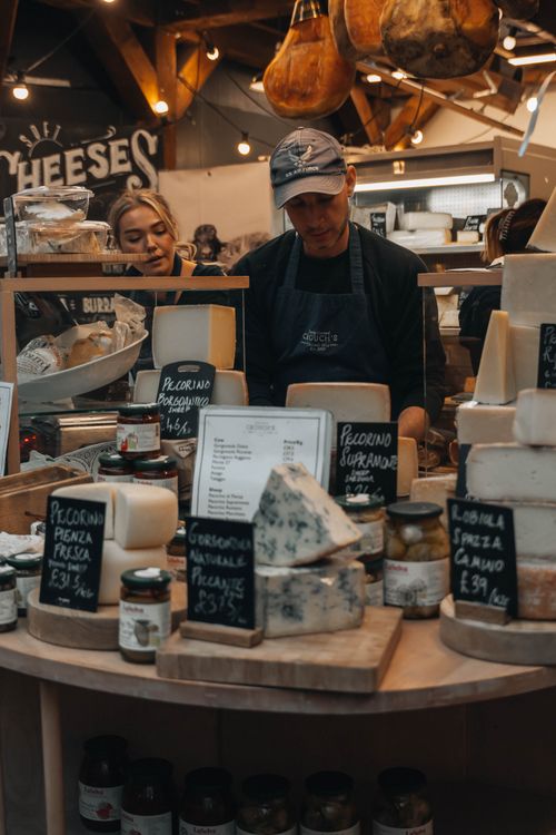 Free Artisan cheese stall in a lively market with diverse cheeses and customers exploring new flavors. Stock Photo