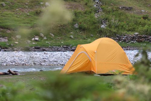 Free Orange tent pitched by a rocky river under a serene sky, surrounded by lush greenery. Stock Photo