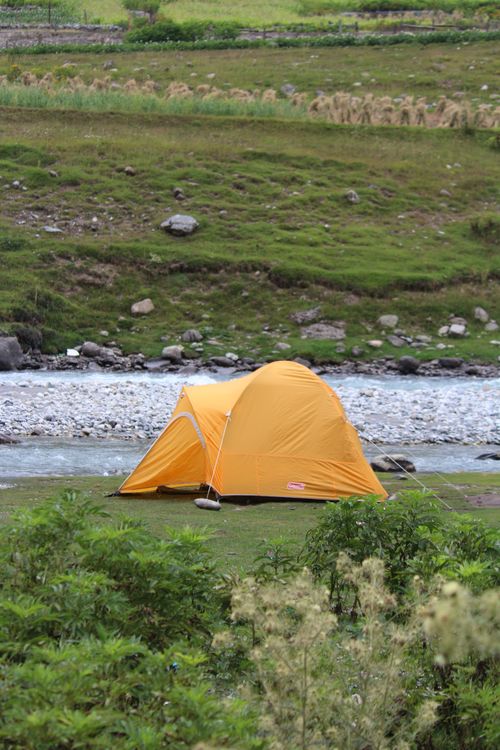Free A serene outdoor camping scene with a yellow tent beside a flowing river in lush greenery. Stock Photo