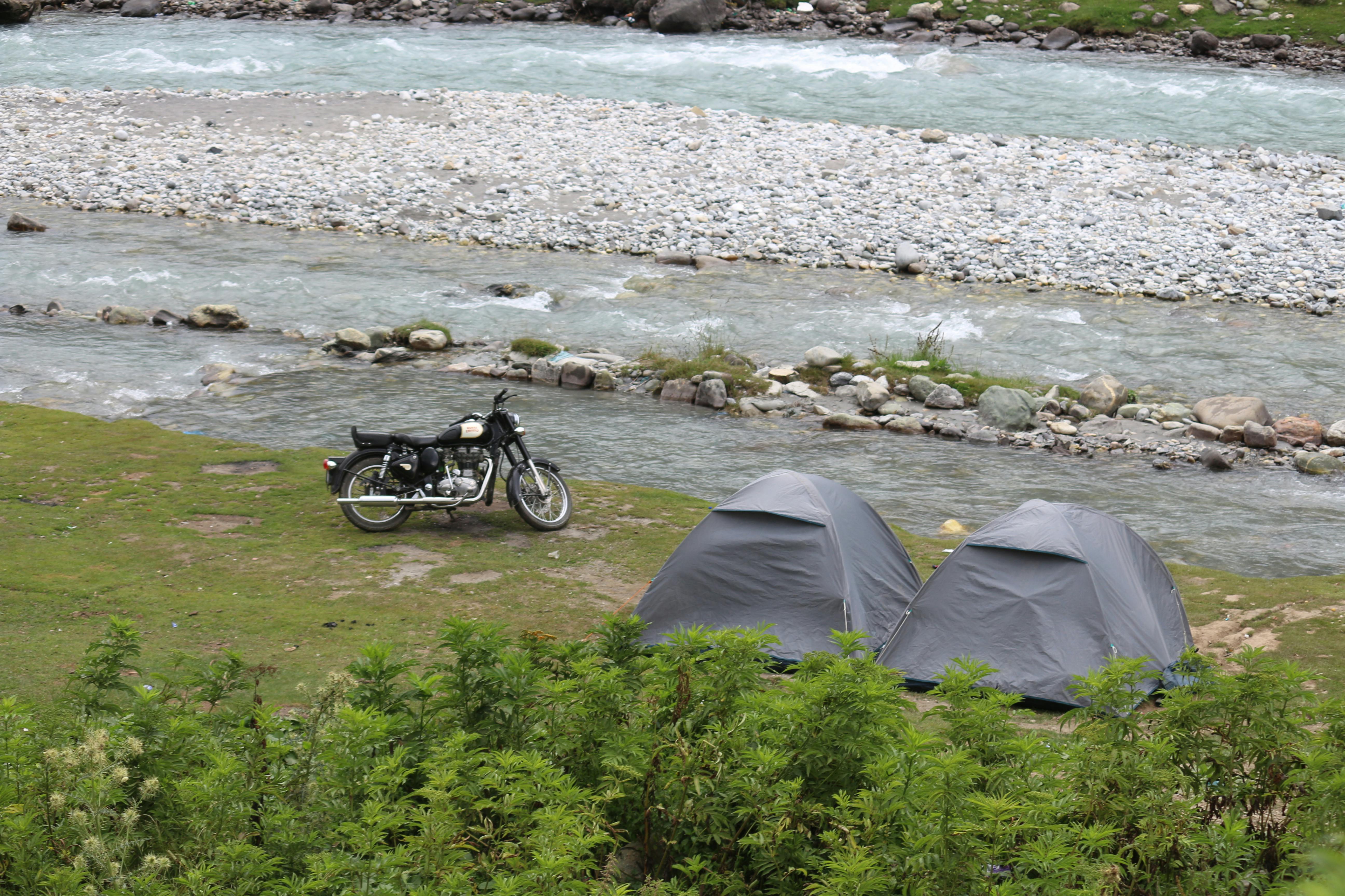 Two tents near a river with a motorcycle on a lush, green campsite.