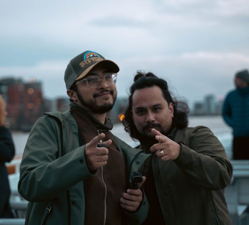 Free Two men smiling and pointing towards the camera during a casual day outdoors. Urban background enhances the lively atmosphere. Stock Photo