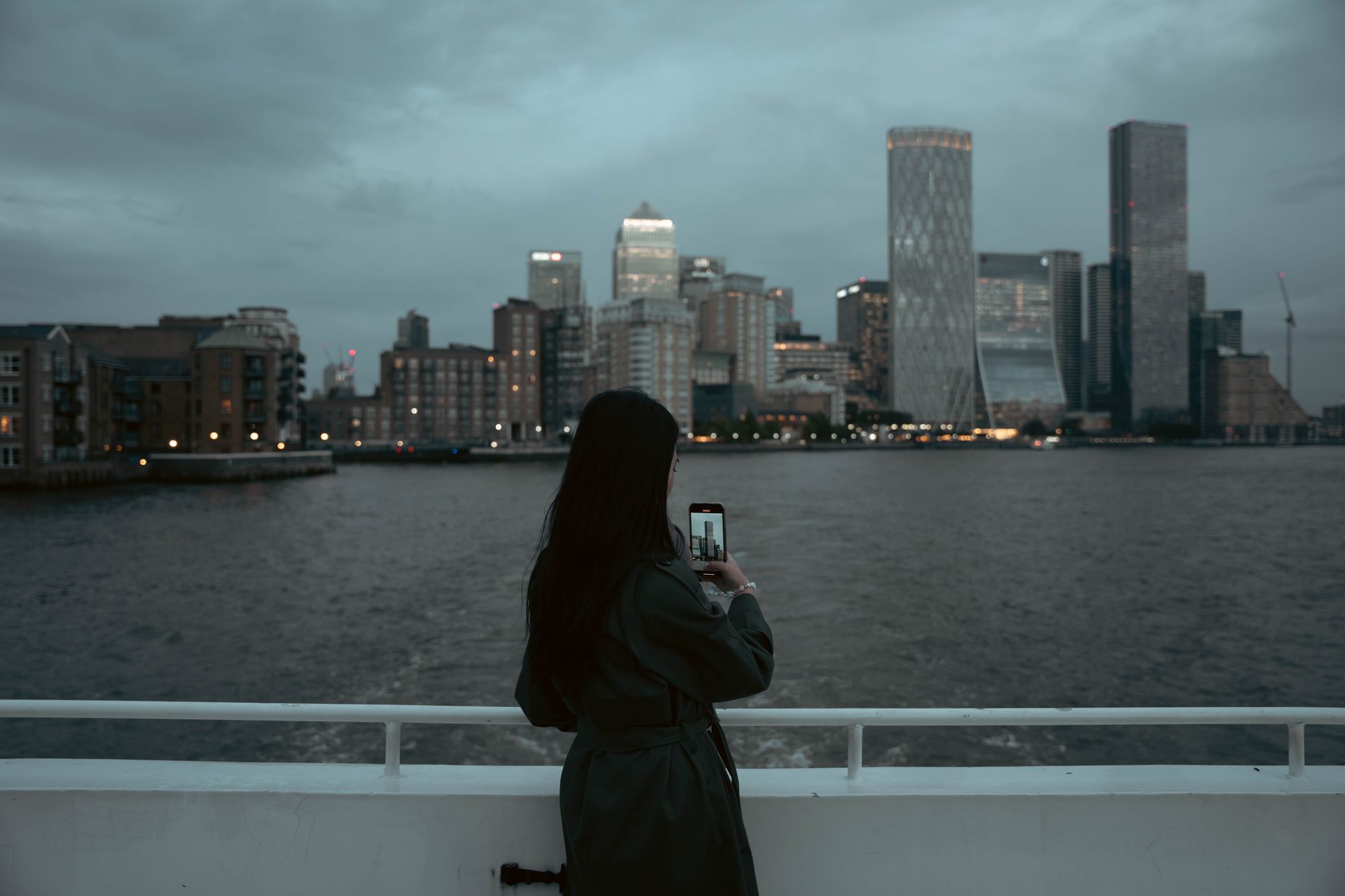 A person photographs the London skyline at dusk with high-rise buildings reflecting in the river.