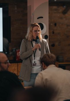A woman speaks to an audience at an indoor event with a microphone in hand.