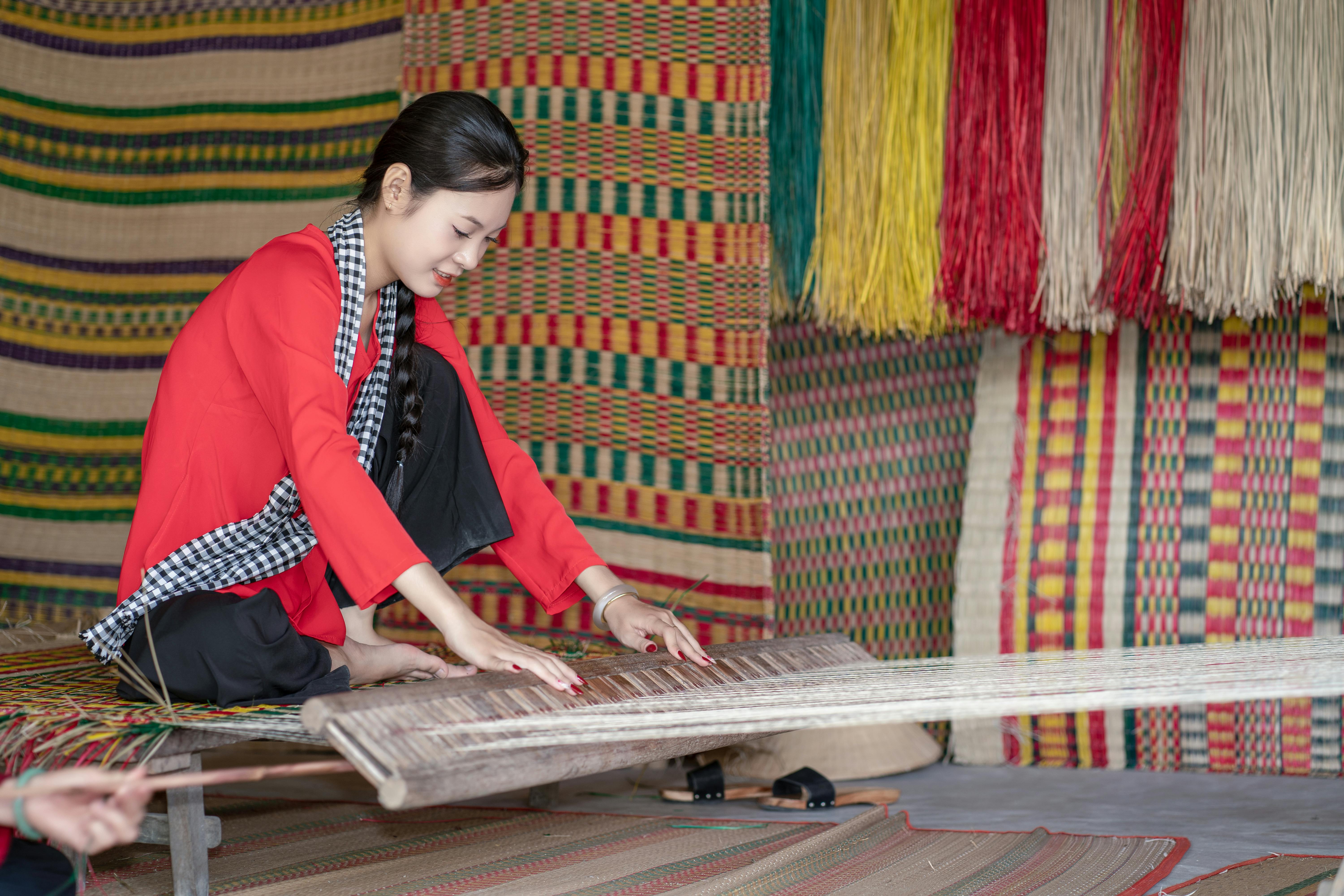 A woman in traditional attire weaving a colorful mat indoors.