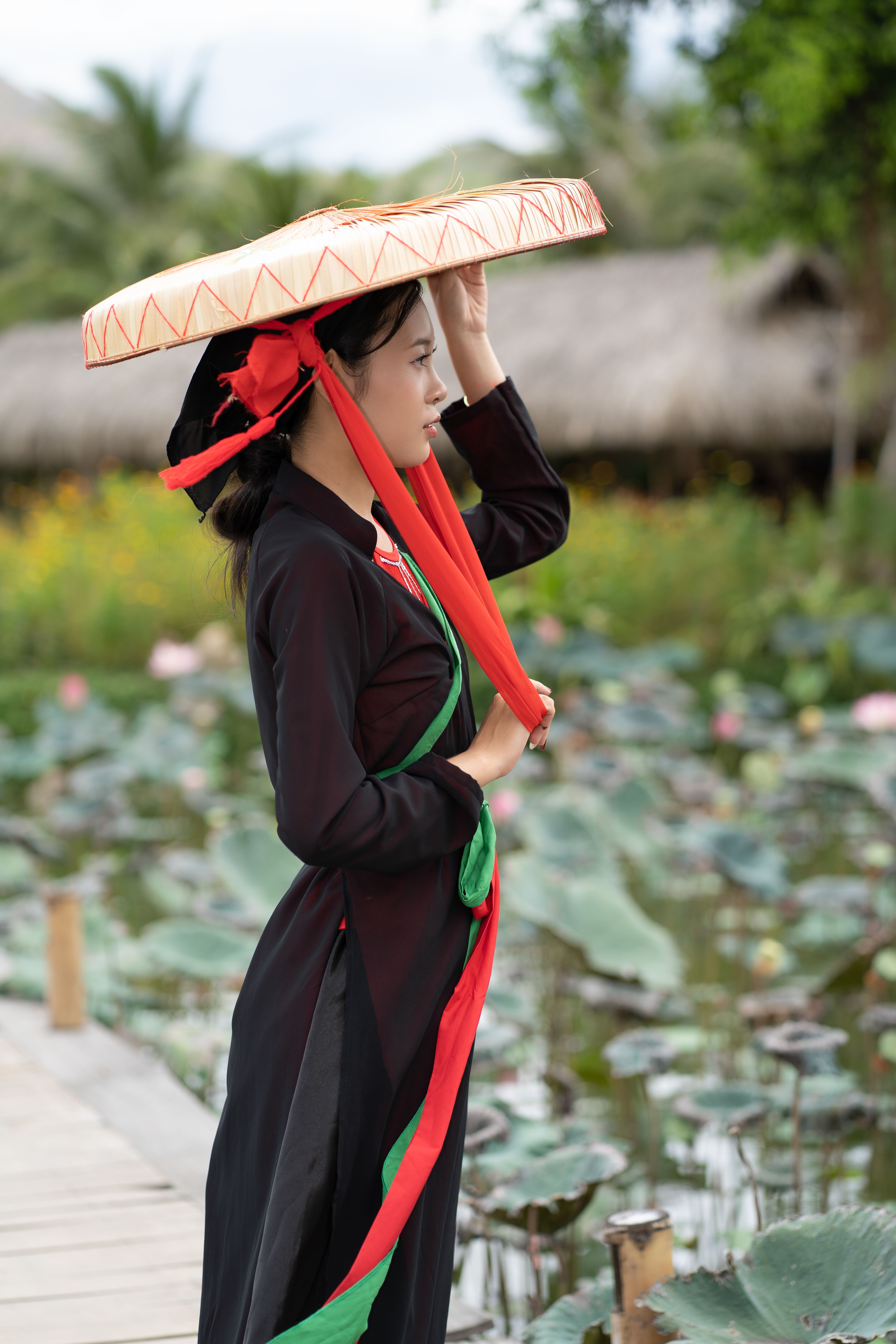 A woman wearing traditional attire stands by a lotus pond, holding a wide-brimmed hat.
