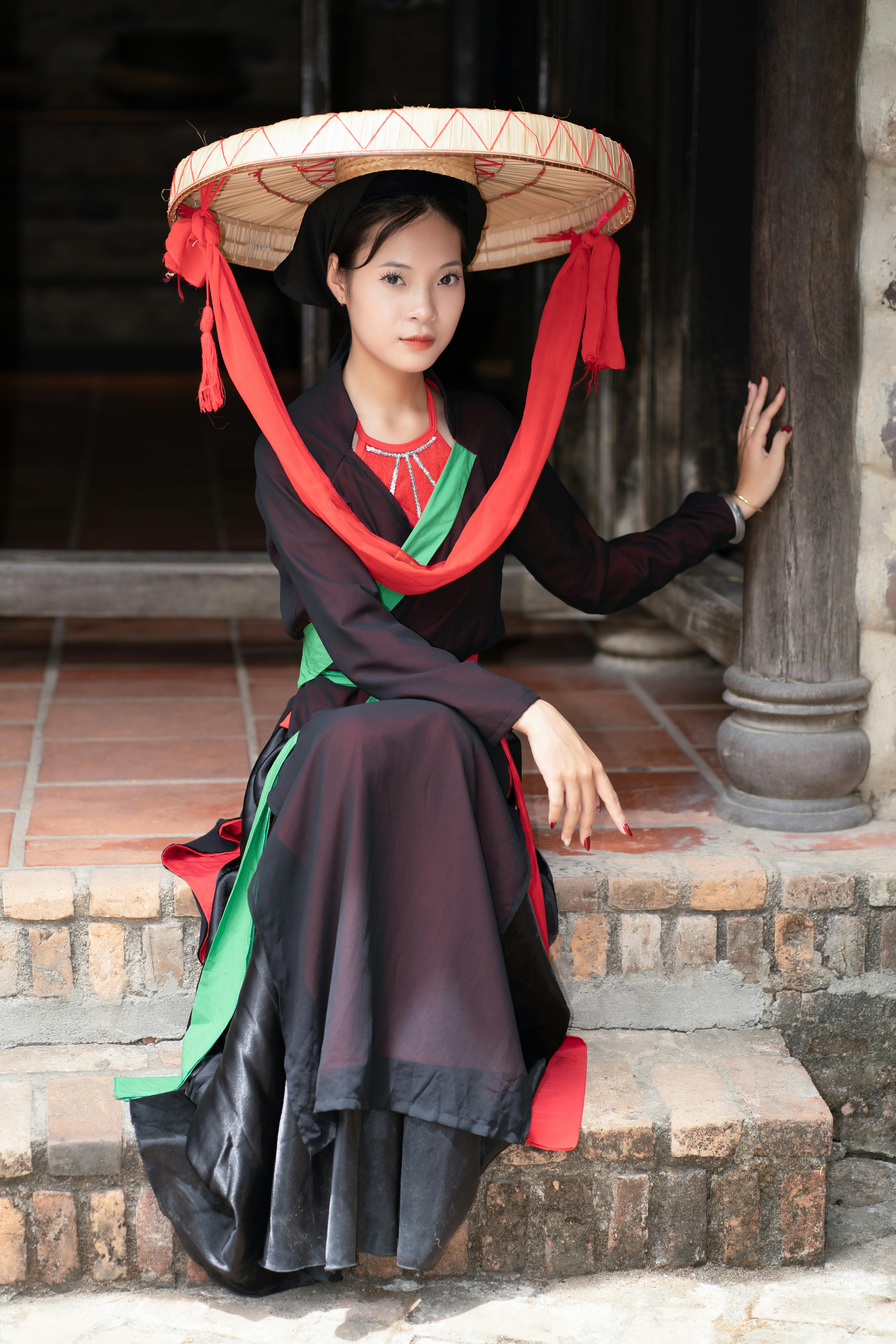 Young woman in traditional Vietnamese dress with conical hat sitting on steps indoors.