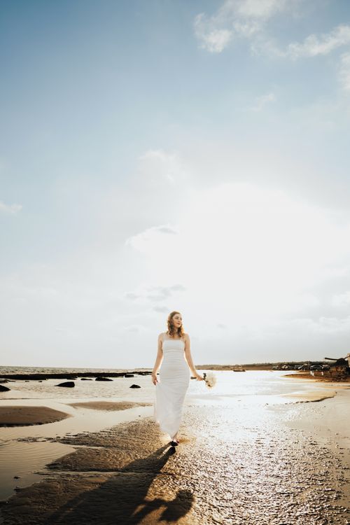 Free A bride in a white dress walks along a sunlit beach, creating an elegant and serene scene. Stock Photo