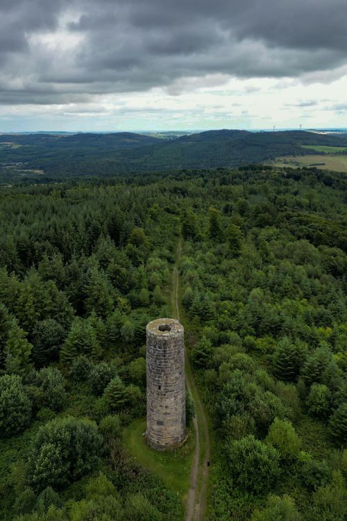 Free Aerial shot of a historic stone tower surrounded by dense green forest under a cloudy sky. Stock Photo