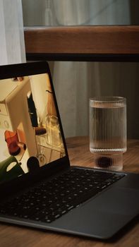 A laptop on a wooden table beside a glass of water in a cozy indoor setting.