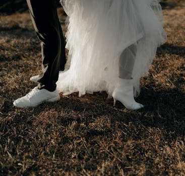 Bride and groom's feet in stylish shoes on grassy field during golden hour.