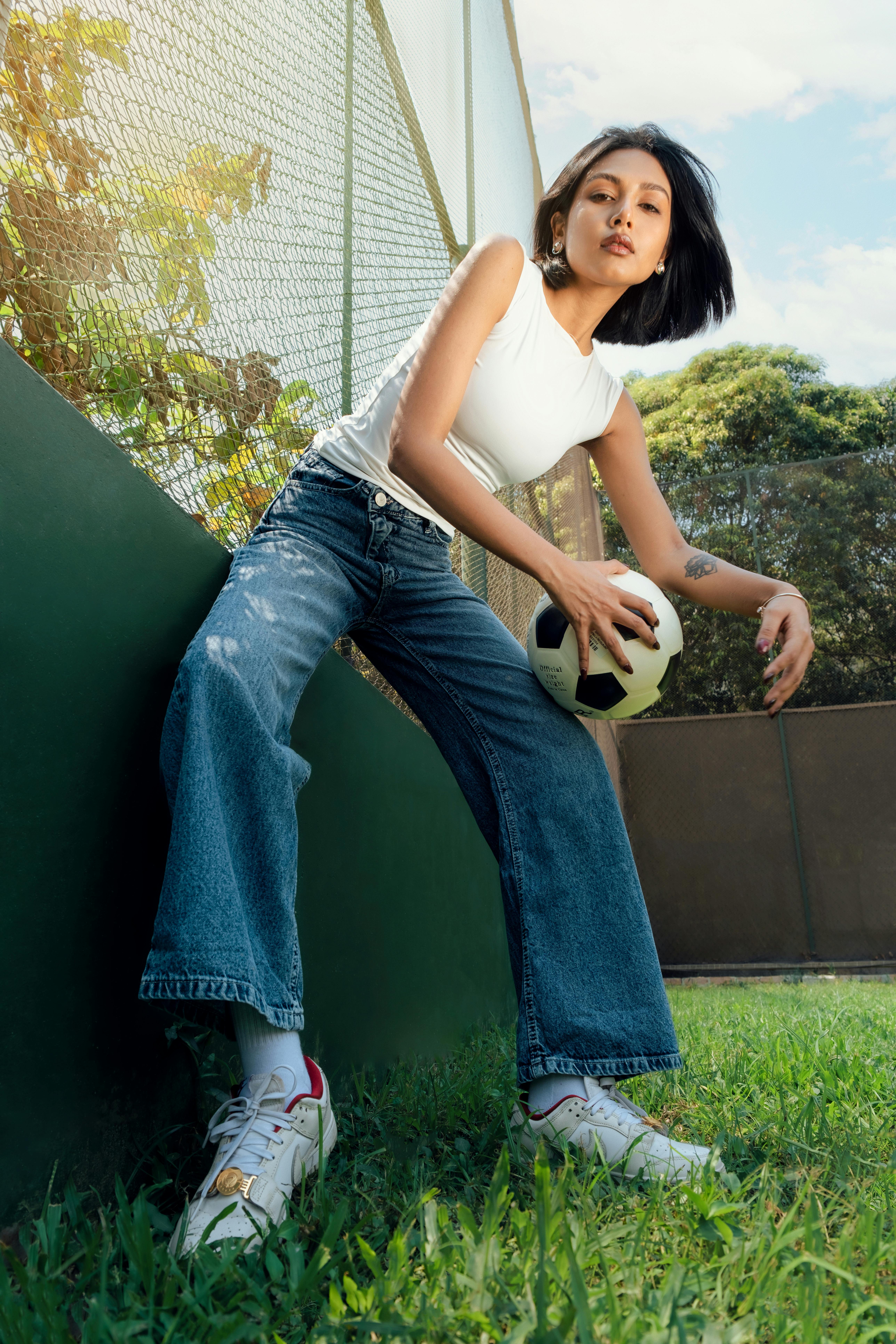 Stylish young woman holding a soccer ball on a sunny day by a sports field.