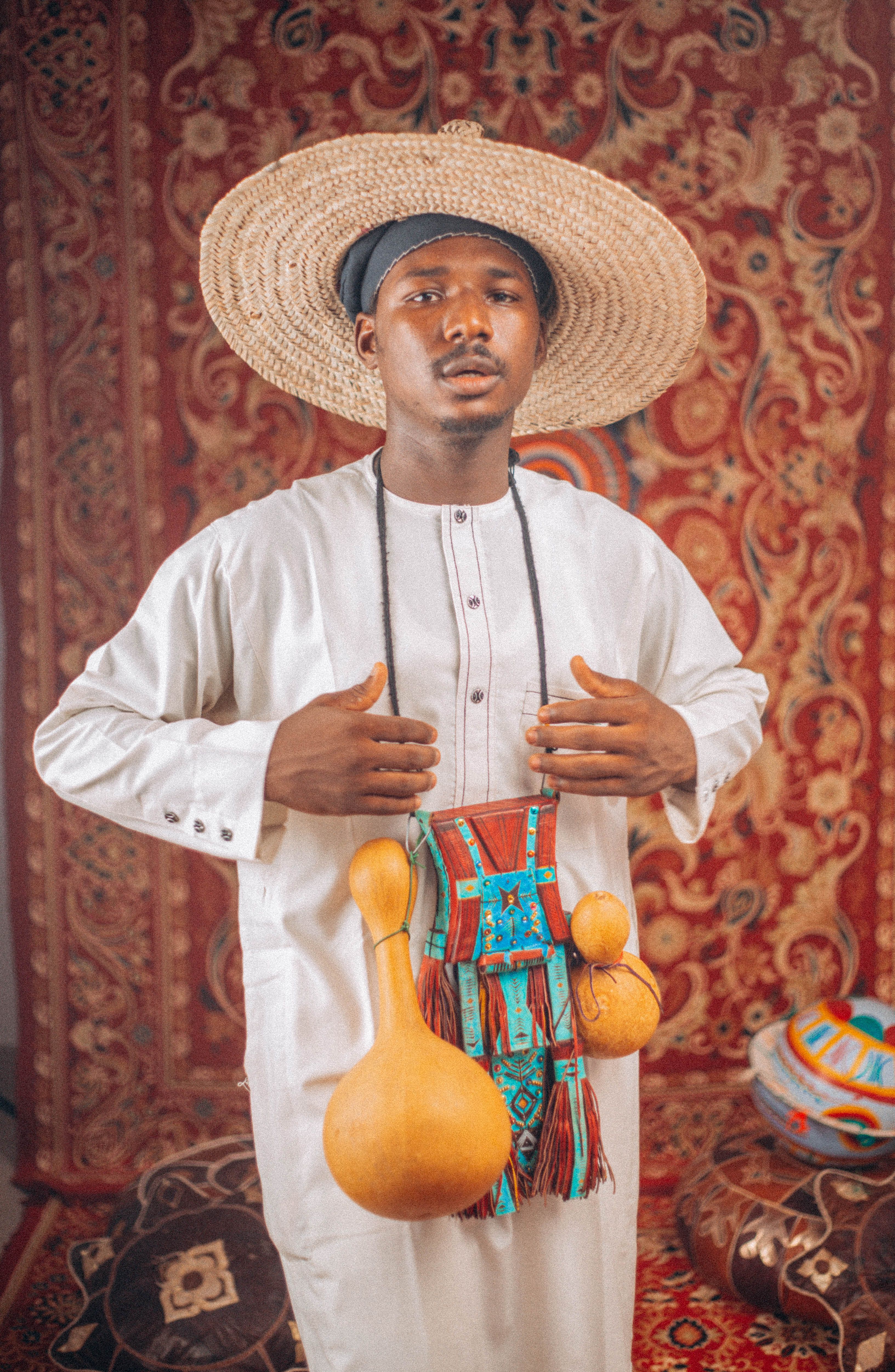 Man in traditional African attire with straw hat, holding decorated gourds, against ornate backdrop.