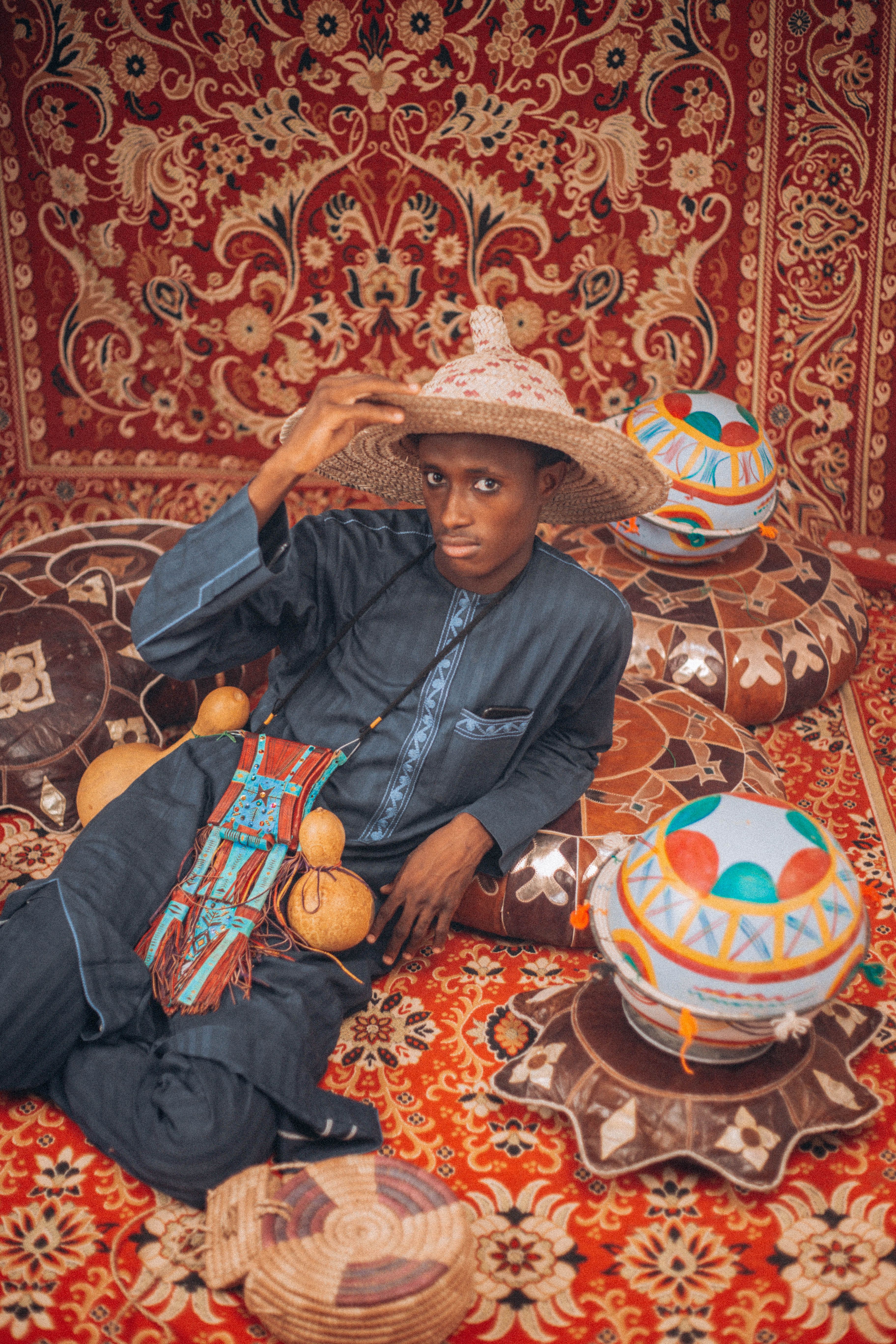 A man in traditional attire sits amidst colorful, patterned cushions and carpet.