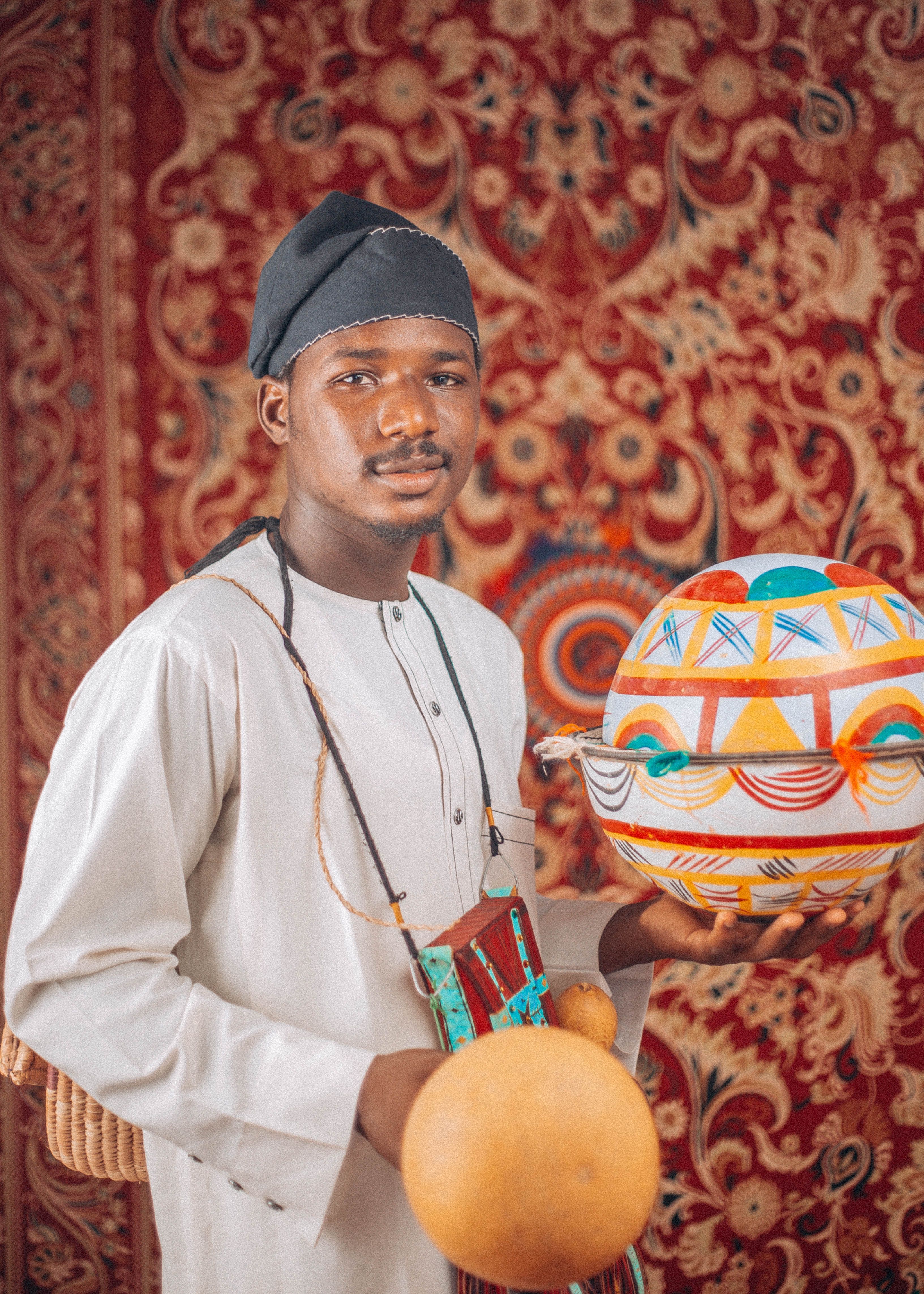 Man in traditional African attire holding colorful tribal pots against a vibrant tapestry.