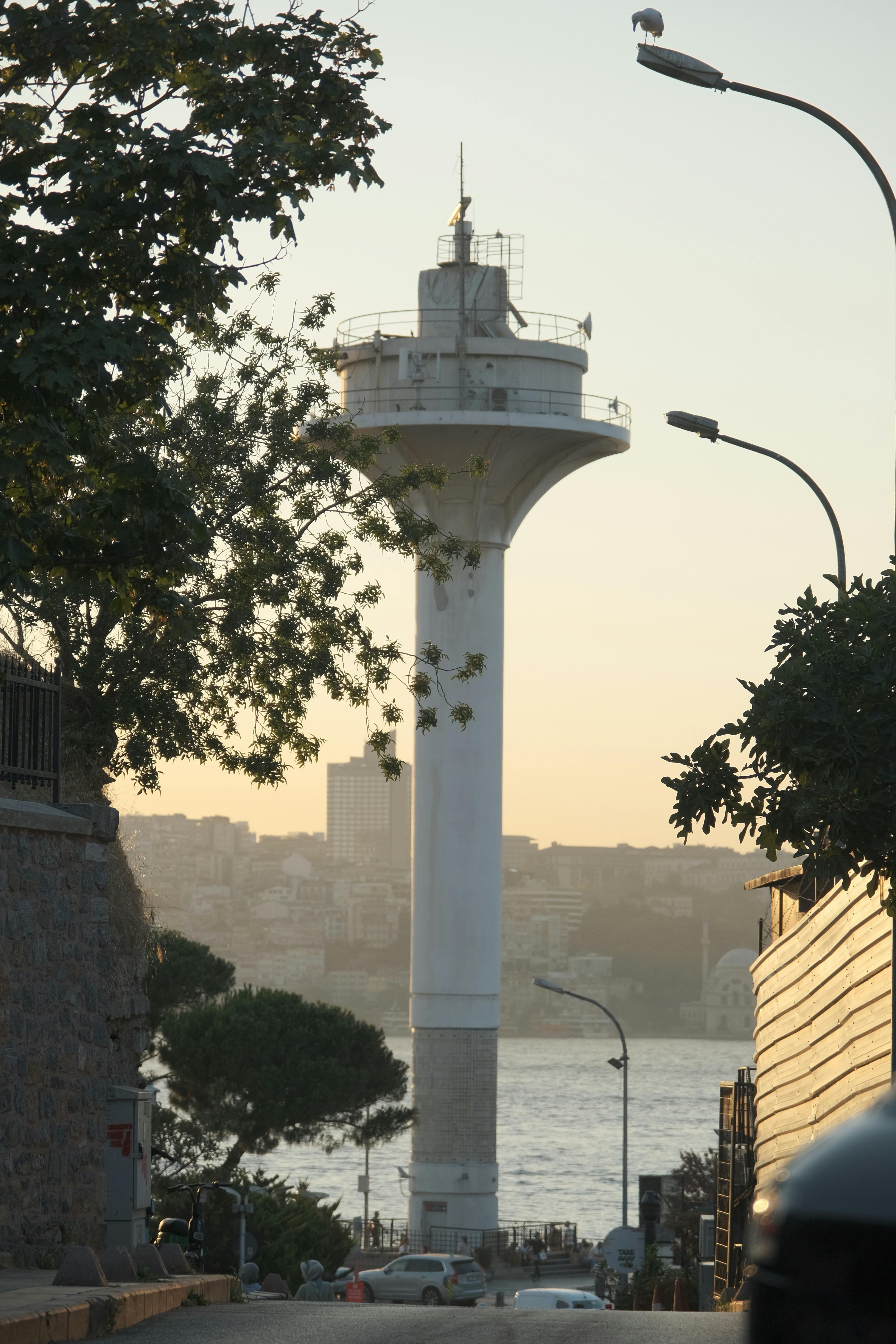 View Of A Towering Lighthouse Near The Sea During Dusk With Trees And Street Lamps.