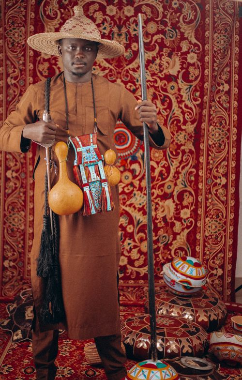Free Man wearing traditional attire holding gourds and staff in a richly decorated room. Stock Photo