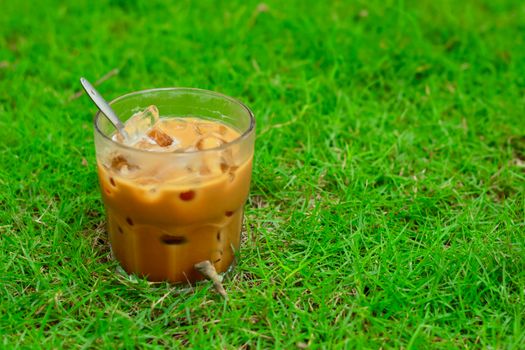 Glass of iced coffee with a spoon, resting on vibrant green grass outdoors.