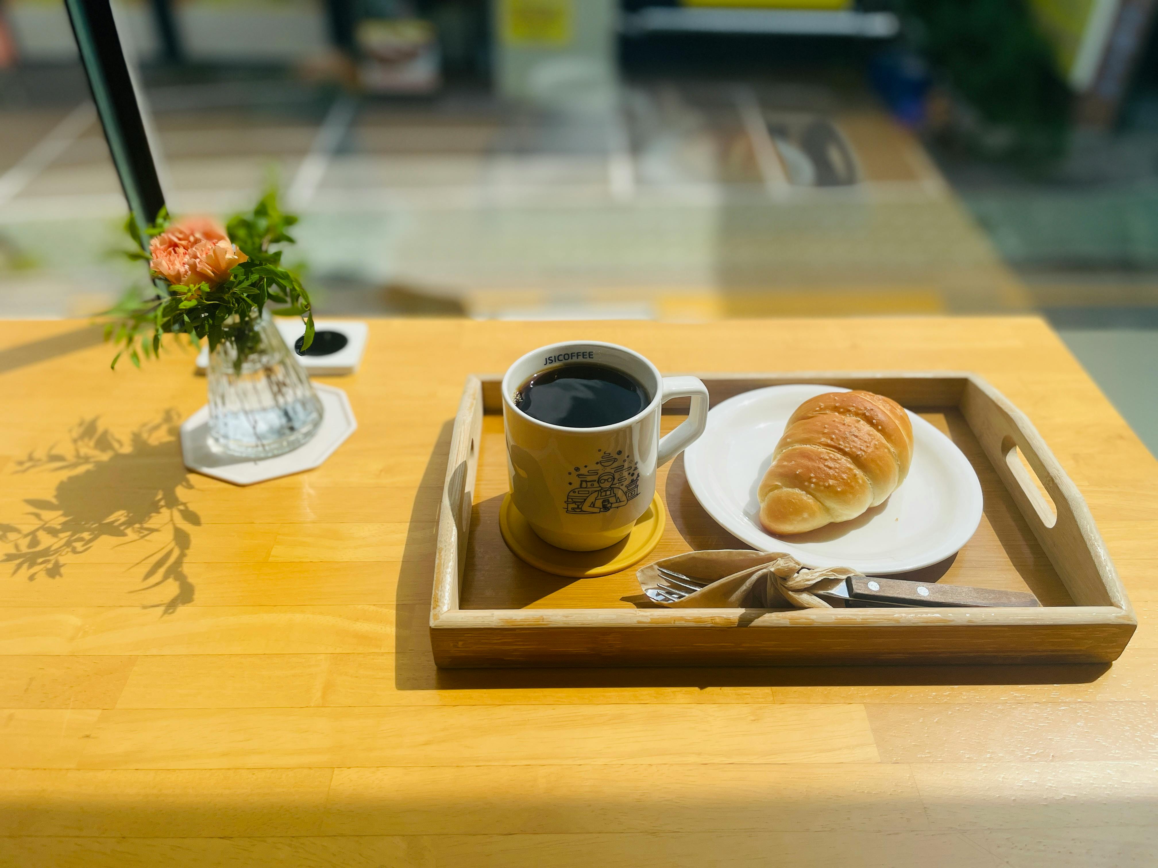 A warm, inviting café setting featuring coffee, a croissant, and a flower vase on a sunny wooden table.