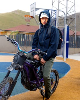 A young man in a hoodie with a bike at an outdoor basketball court surrounded by hills.