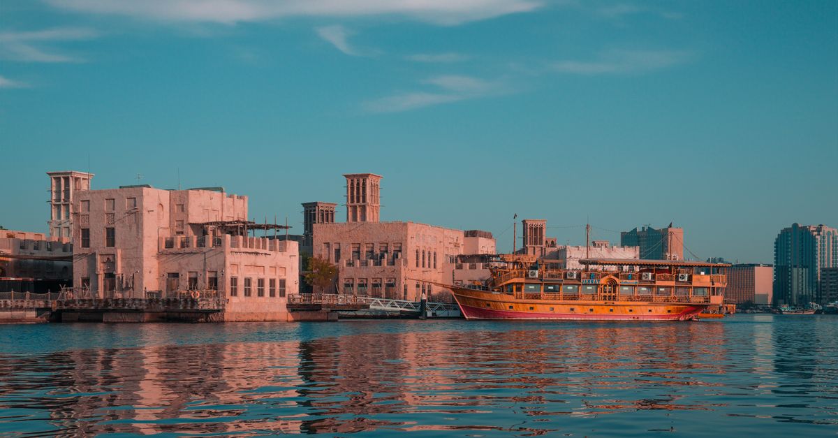 Scenic view of a traditional dhow boat near historical buildings along Dubai Creek at sunset.