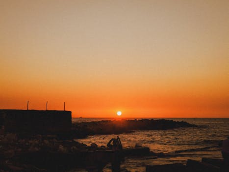 Tranquil sunset scene over the ocean with silhouetted figures along the rocky coast.