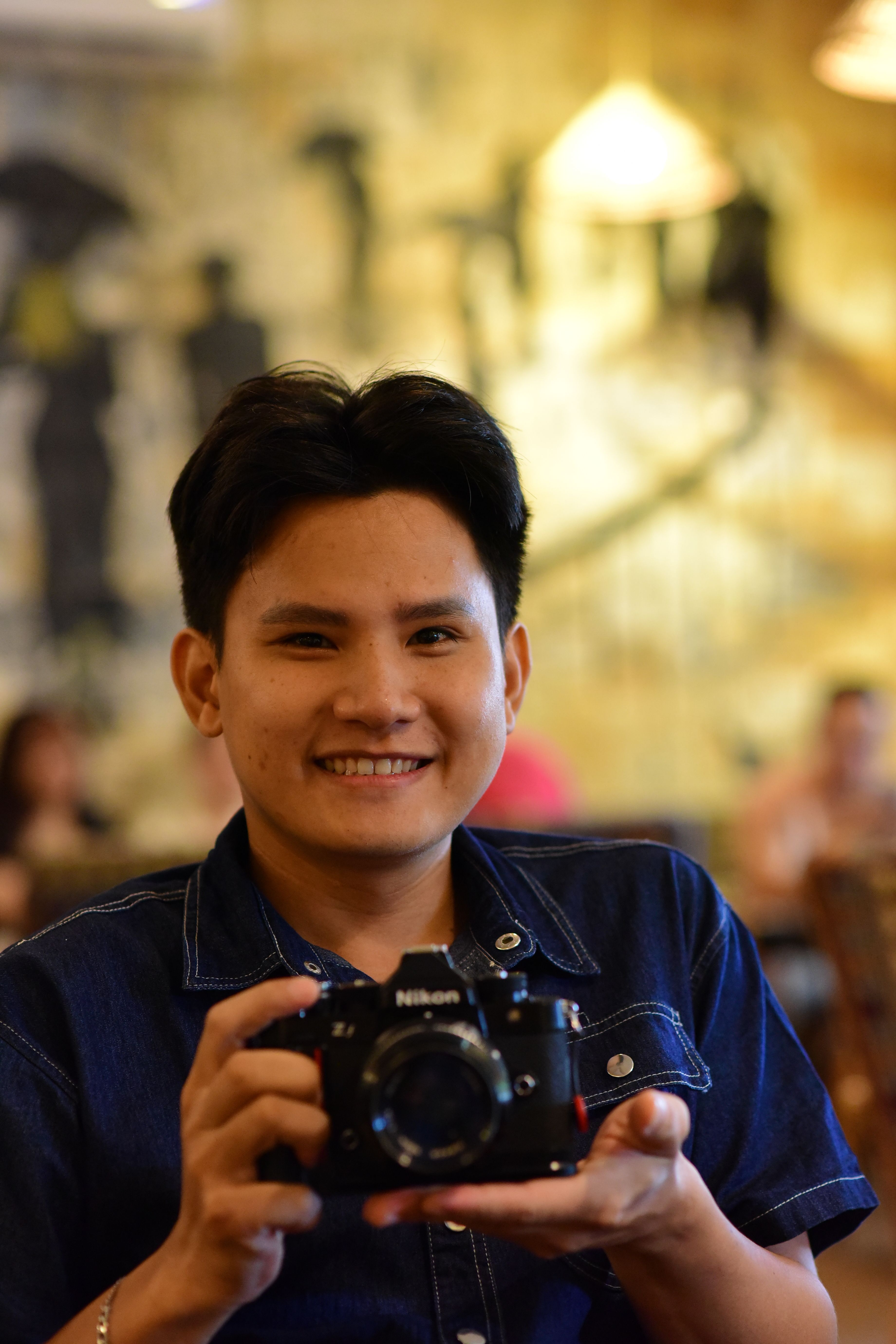 Cheerful person posing with a Nikon camera inside a cozy cafe setting.