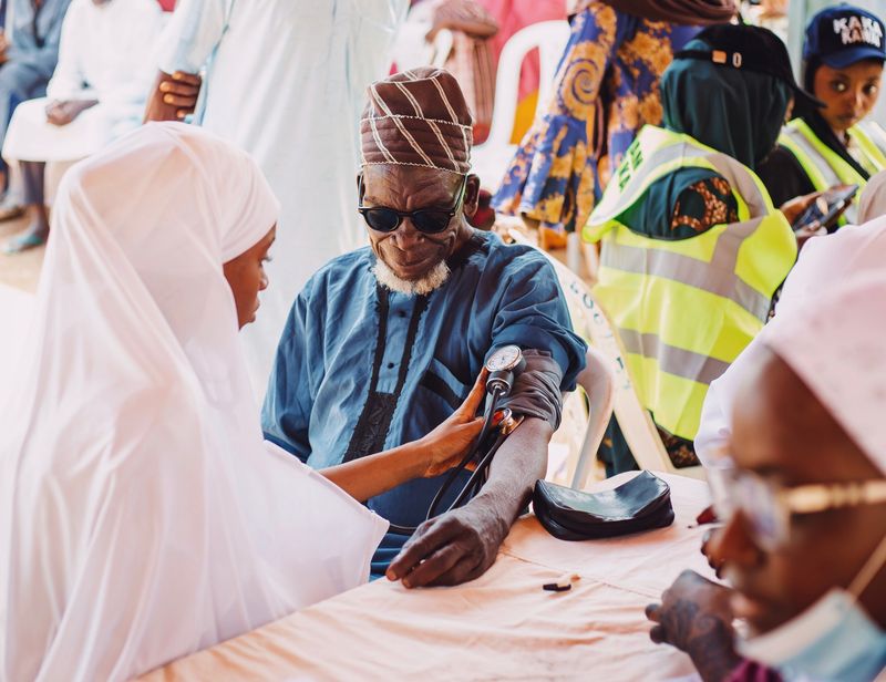 An older Black man in a blue kaftan and embroidered cap has his blood pressure taken with an upper-arm cuff by a health worker in a white headscarf at a community health outreach event.