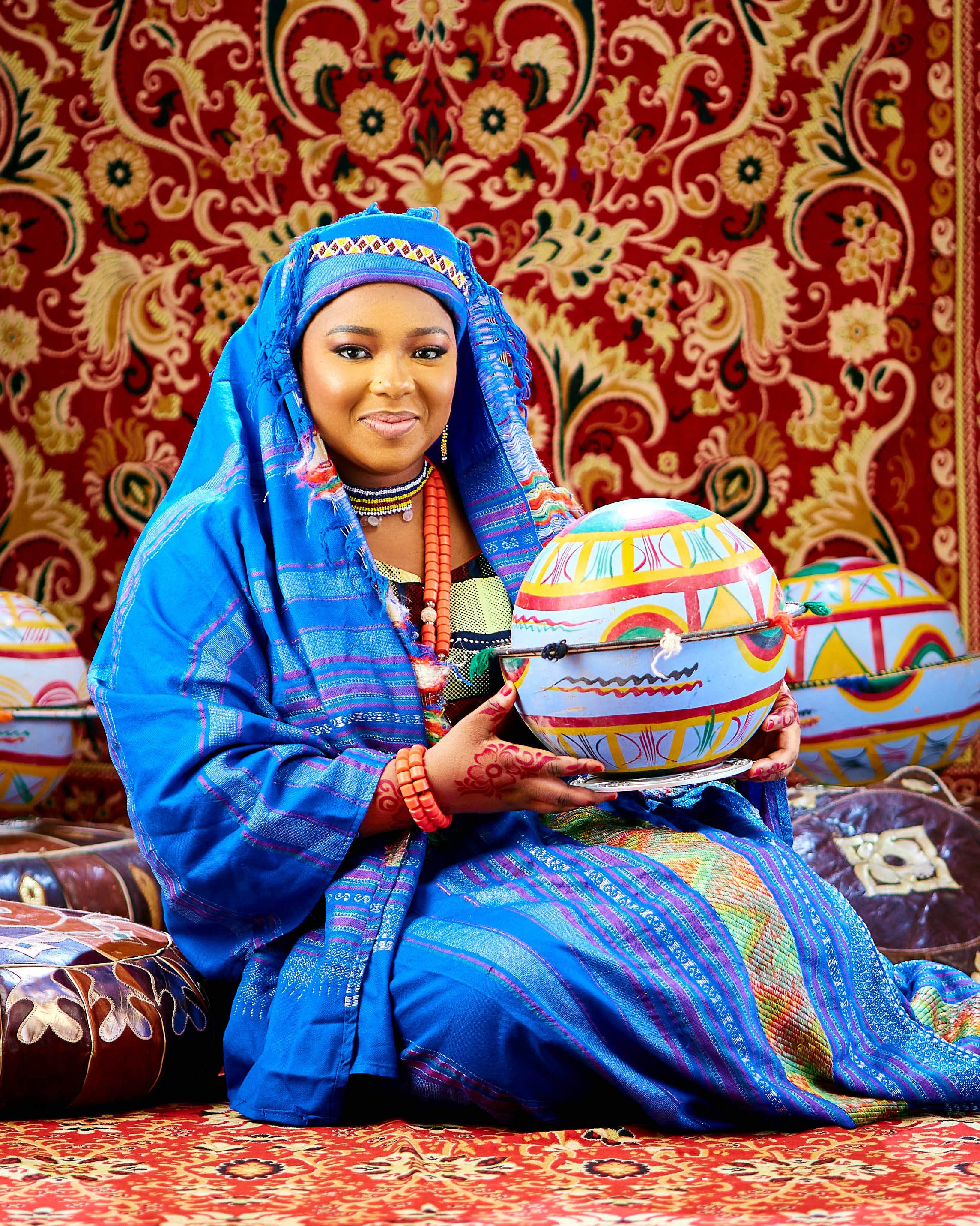 An African woman in vibrant traditional attire holds colorful pottery, surrounded by ornate textiles.