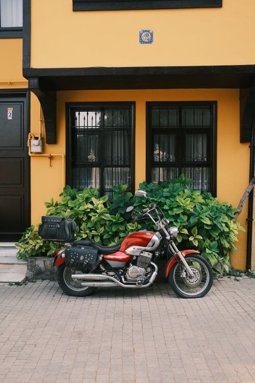 Free Vintage red motorcycle parked in front of a yellow building with green plants. Stock Photo