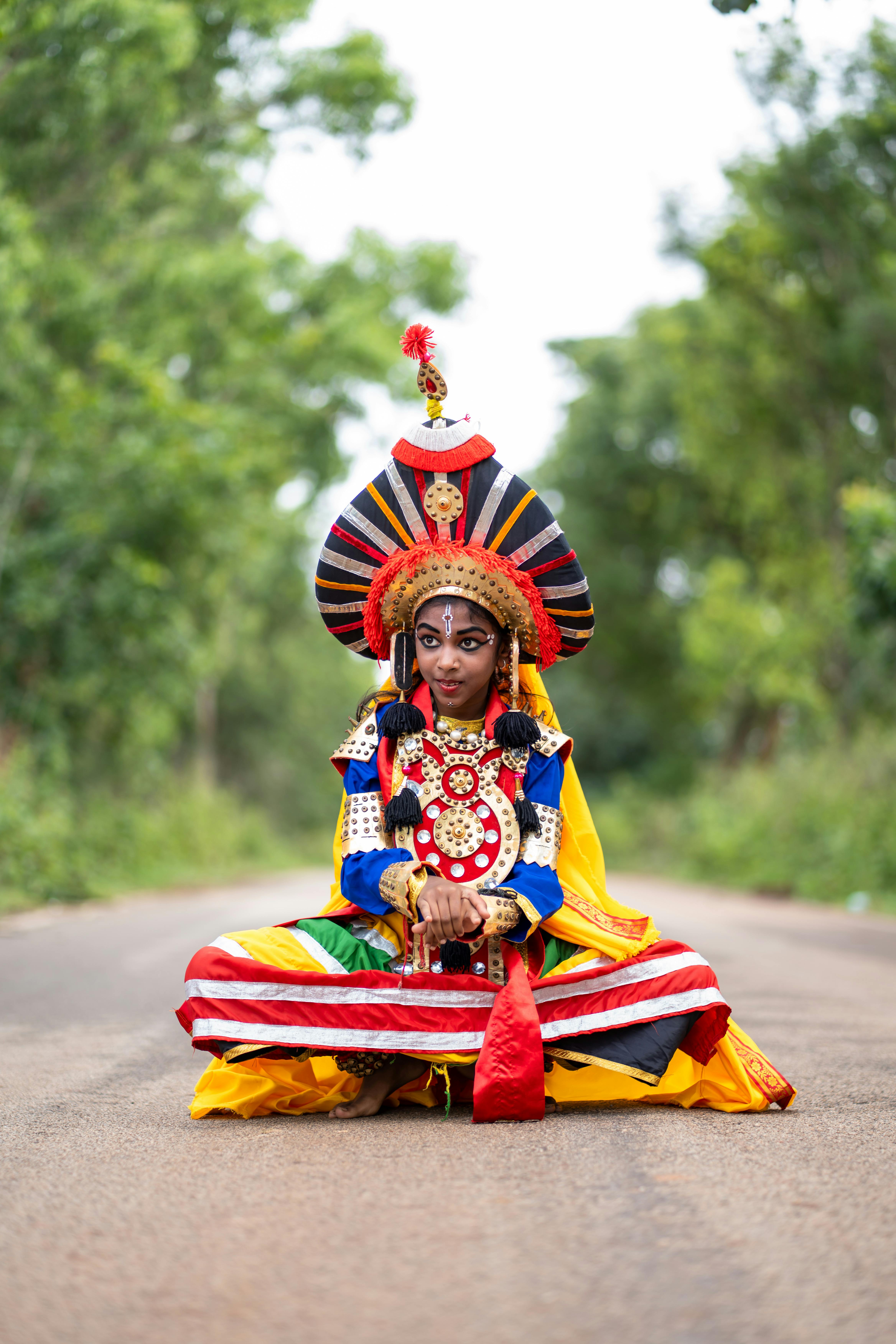Colorful traditional dancer in a vibrant costume seated on a road with greenery.