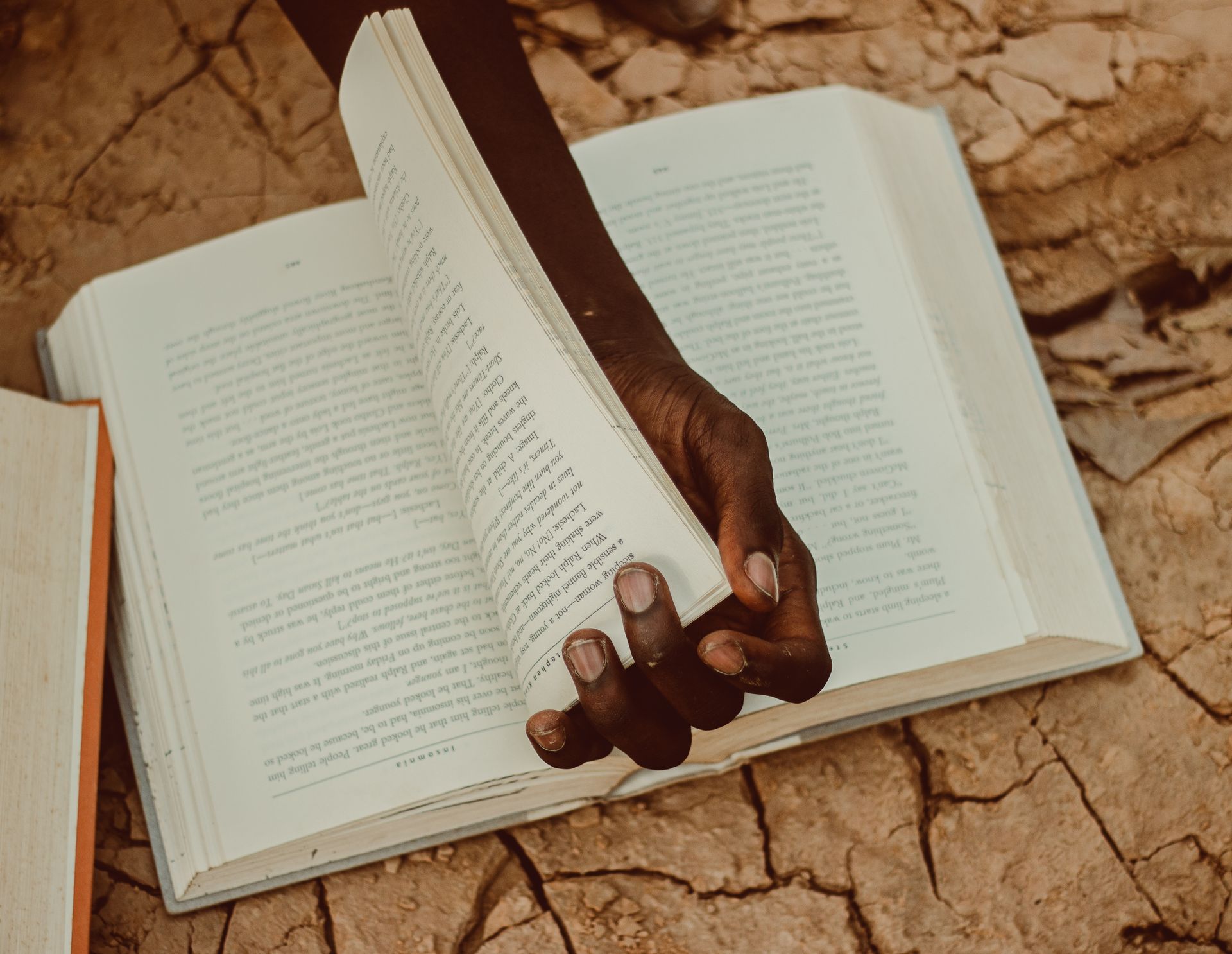A person holds a book open on parched, cracked earth, symbolizing knowledge amidst adversity.