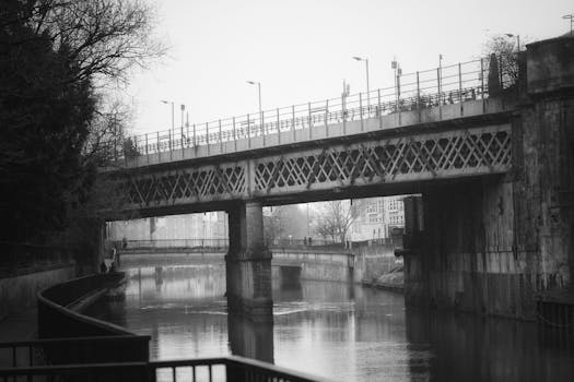 A monochrome view of a railway bridge crossing a calm river in an urban setting.