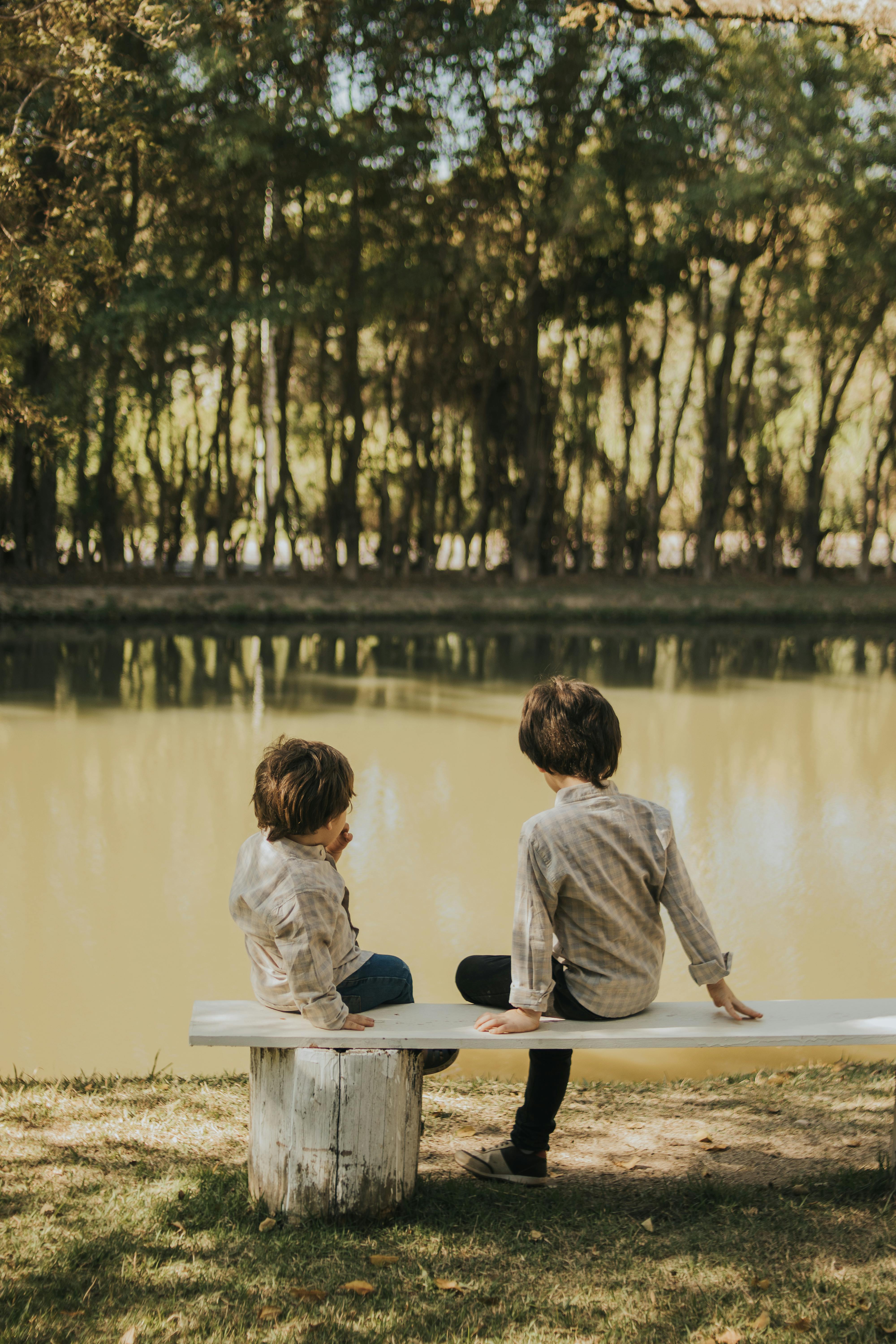 Two kids sitting on a bench by a serene lake in a wooded area, enjoying the peaceful outdoors.