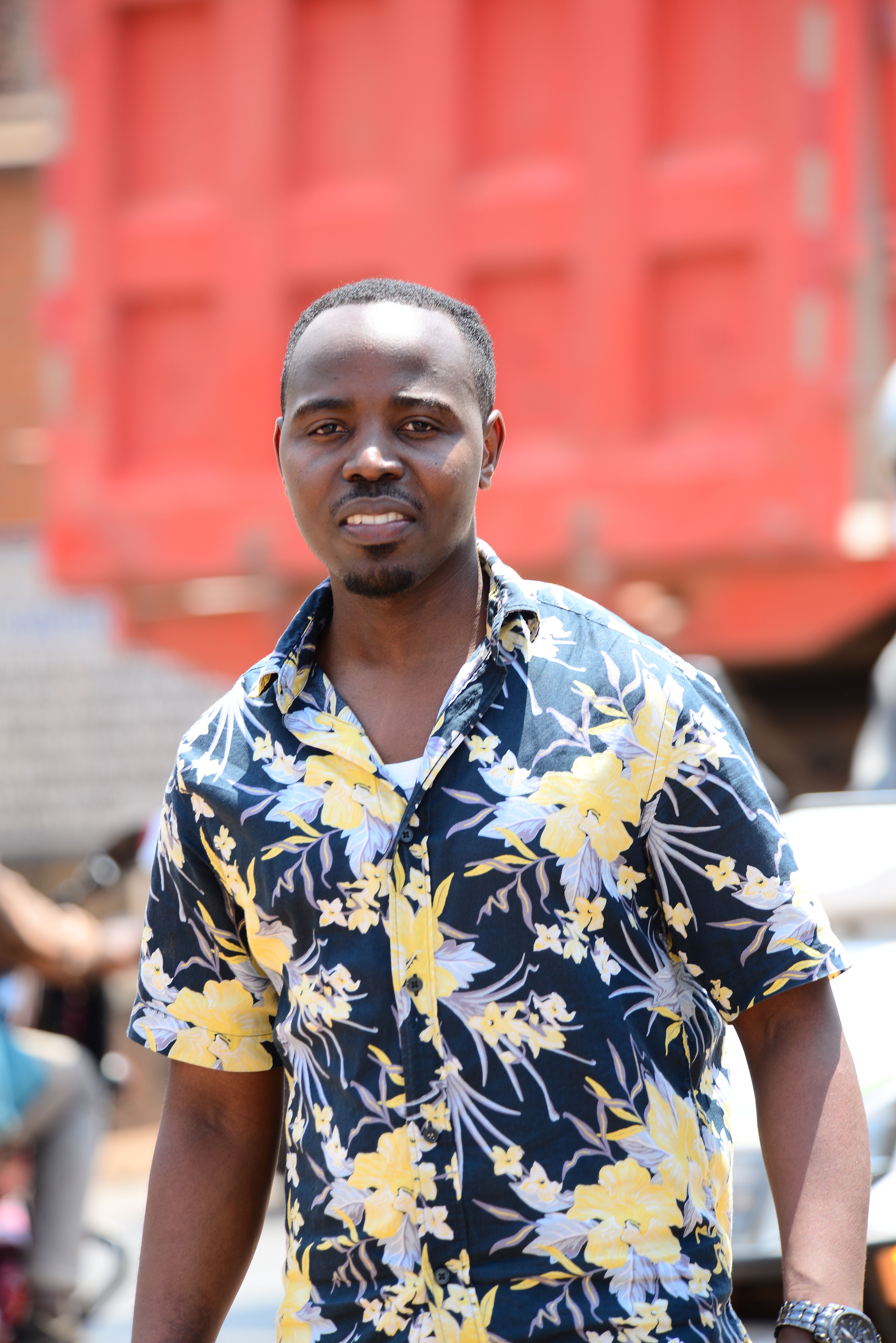 A cheerful man in a floral shirt smiling outside on a sunny day with a red truck backdrop.