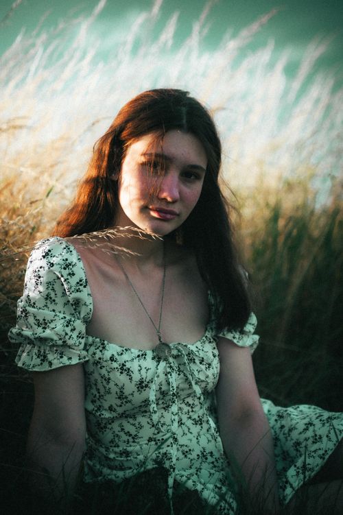Free A young woman in a patterned dress sits in a golden field, captured during sunset for a moody aesthetic. Stock Photo