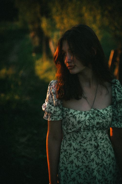 Free A young woman stands in a floral dress, illuminated by warm sunset light in a serene outdoor setting. Stock Photo