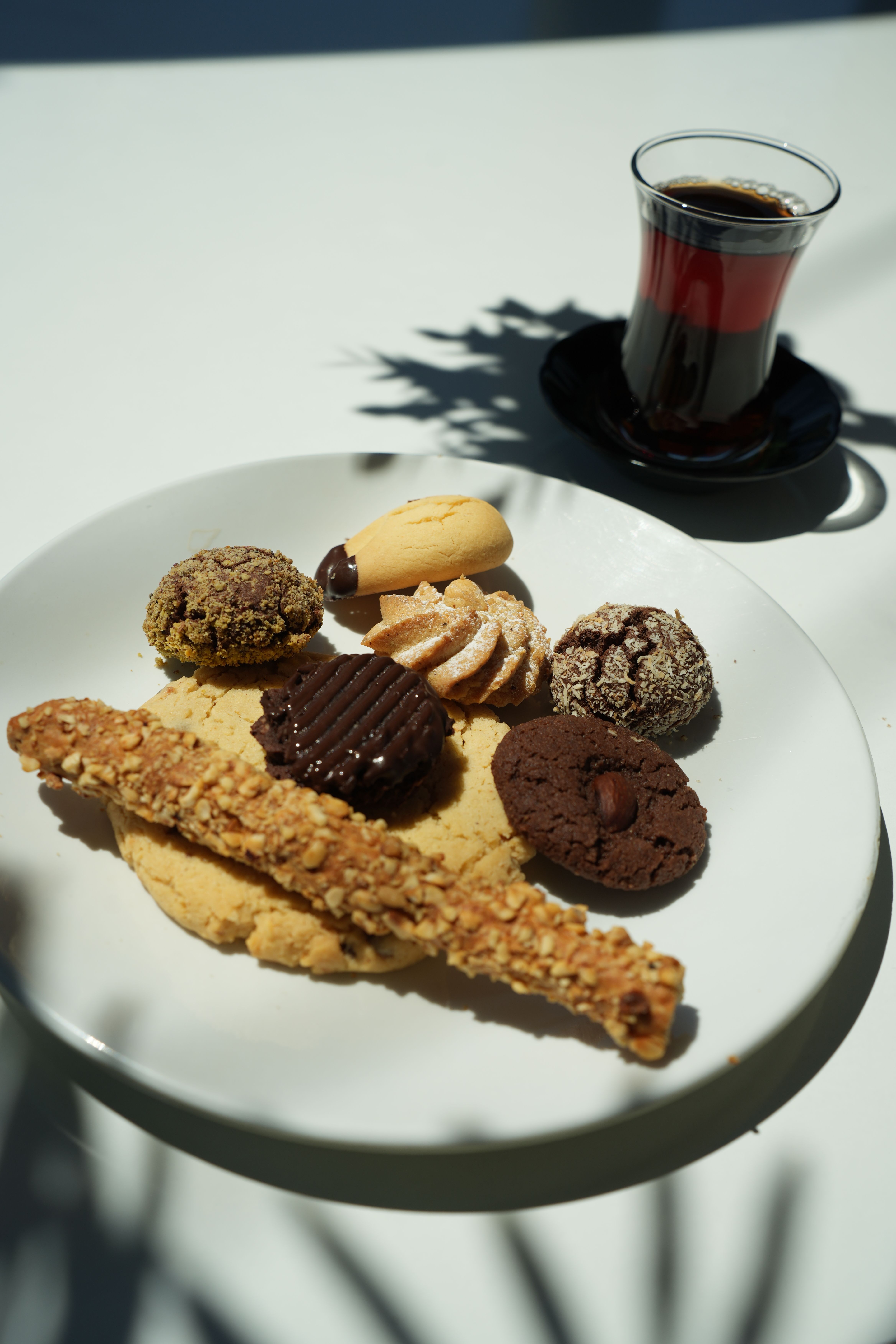 A plate of assorted cookies with a glass of Turkish tea on a sunlit white table.