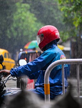 A motorcyclist wearing a red helmet and blue raincoat rides in rainy urban street.