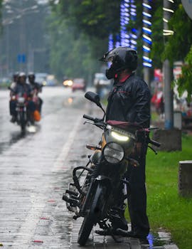 Motorcyclist waits in rain on city street, wearing rain gear, with helmet.